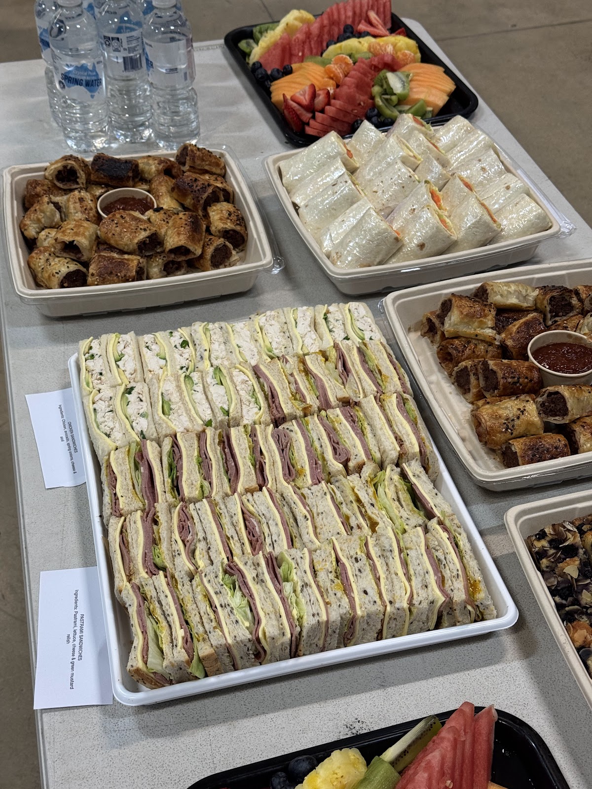 Catering spread of sandwich platters, sausage rolls and fresh fruit